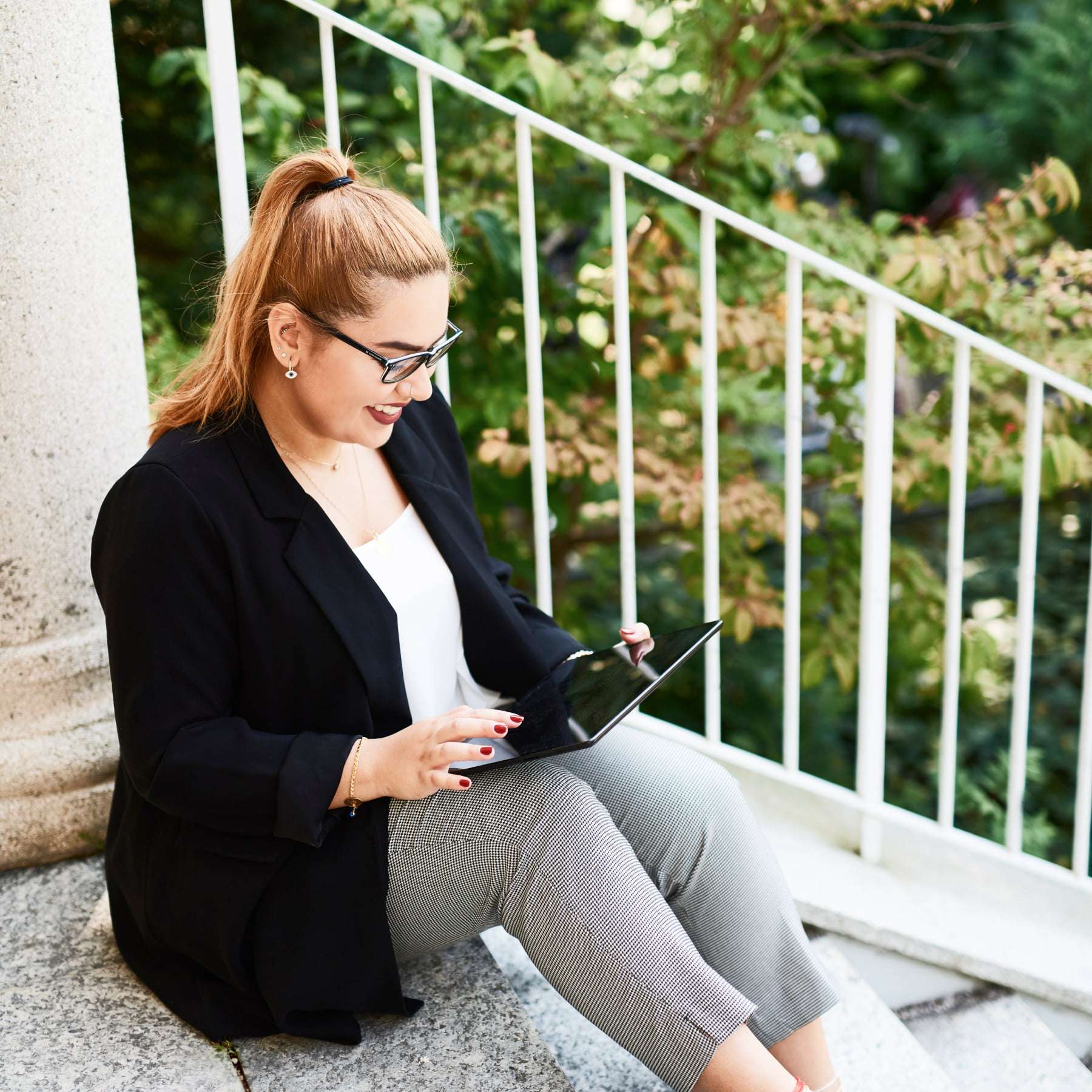 Eine Frau sitzt auf einer Treppe mit weißem Geländer vor Bäumen und hält ein Tablet in der Hand, das sie bedient.