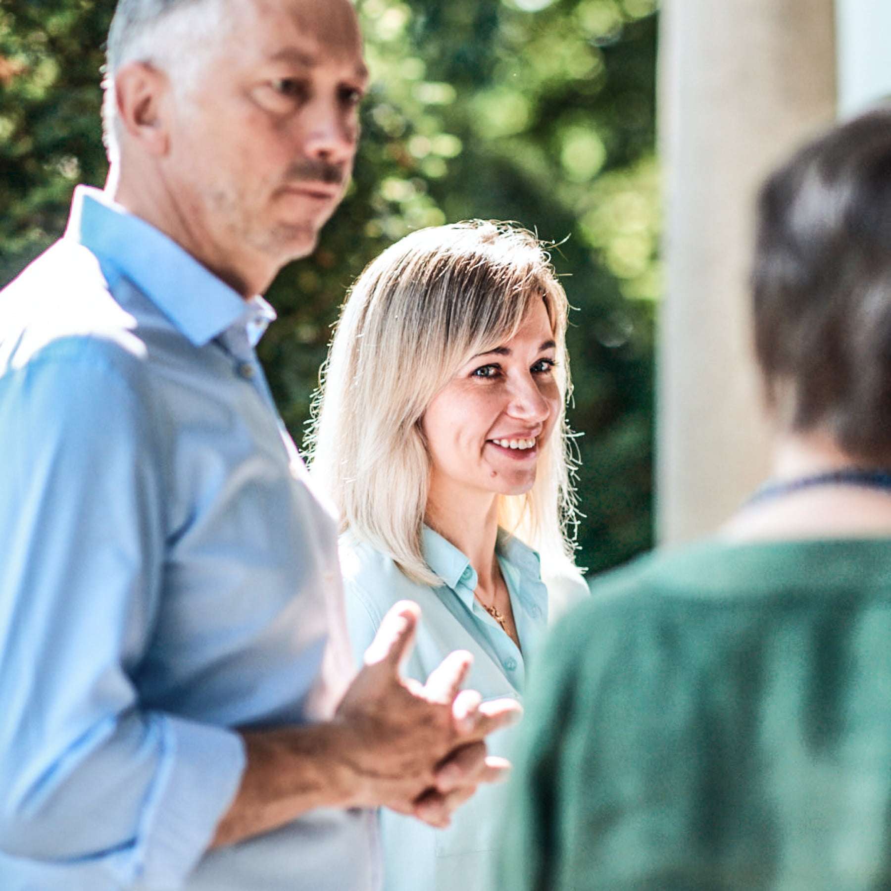 Ein Mann und zwei Frauen stehen neben einer Säule und vor grünen Bäumen und sind ins Gespräch vertieft