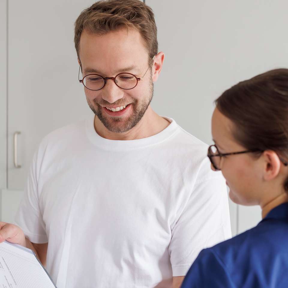 Ein Mann mit Brille und weißem T-Shirt lächelt, während er mit einer Frau in blauer Kleidung spricht und ein Dokument betrachtet.
