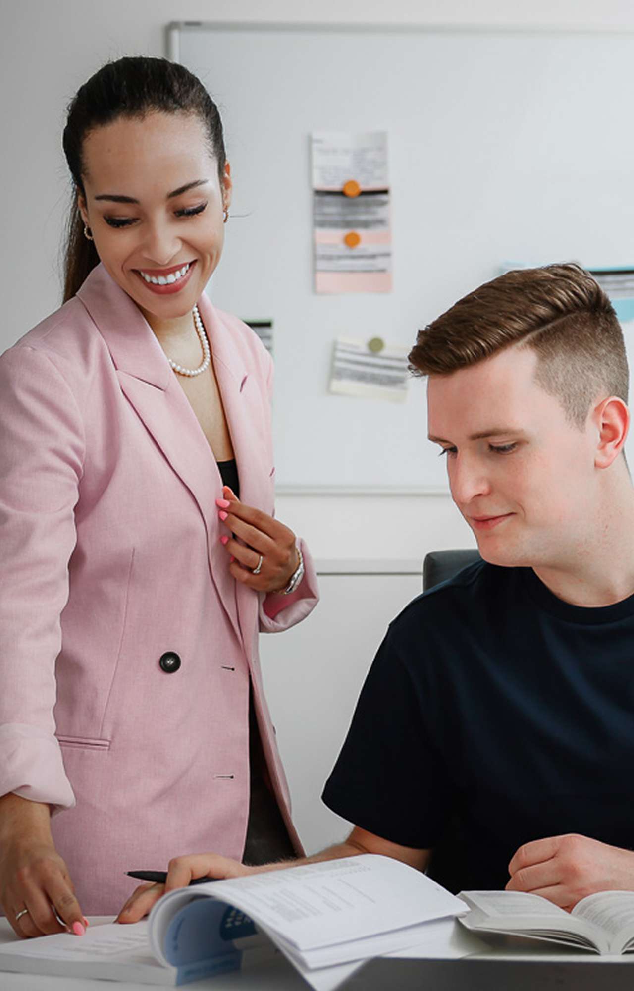 Eine Frau in einem rosa Blazer steht neben einem Mann, der an einem Tisch mit Unterlagen arbeitet.