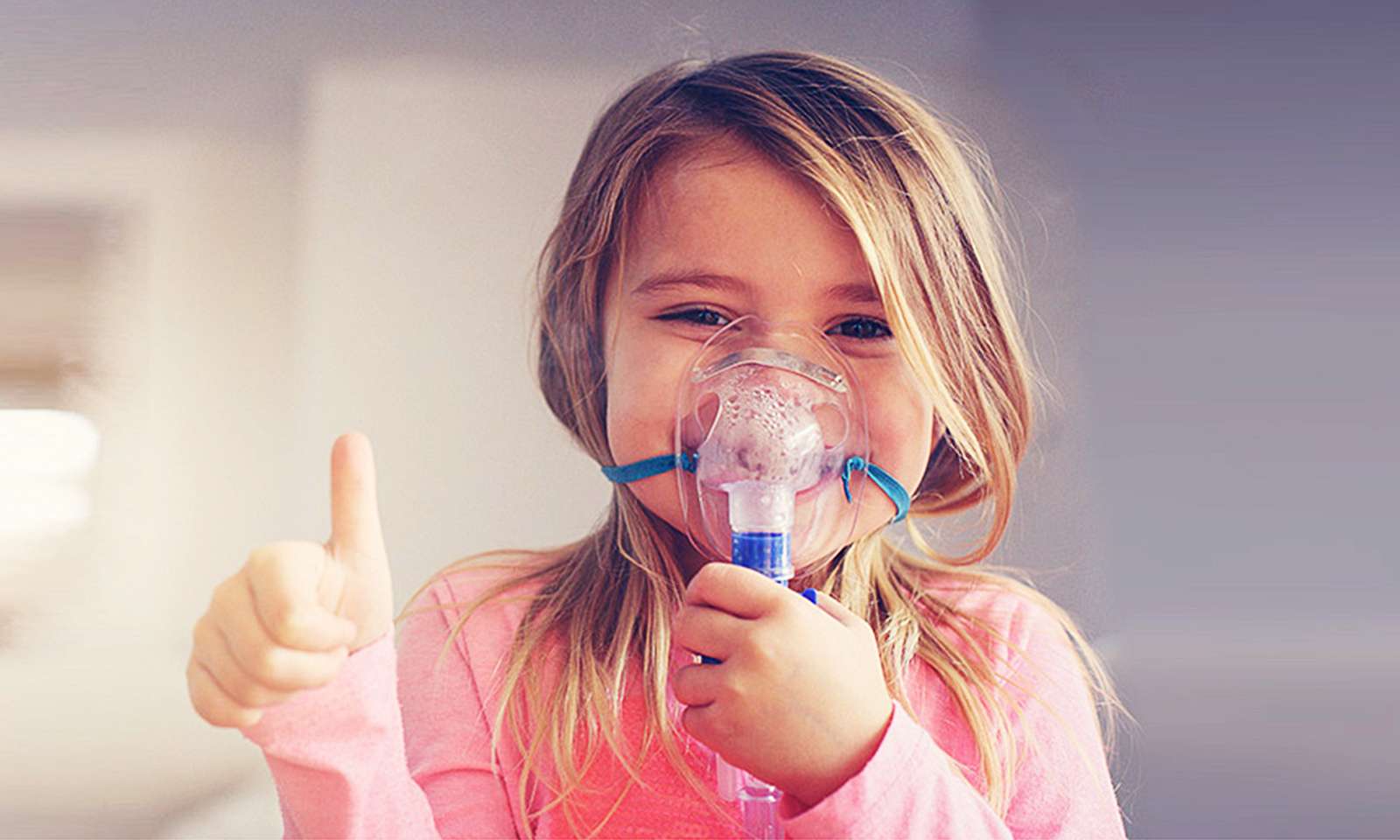 Ca. 4-jähriges Kind mit blauer Inhalationsmaske streckt den Daumen hoch und lacht unter der Maske